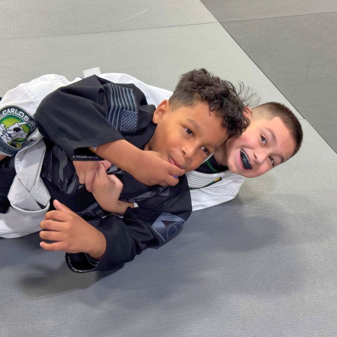 Two children in black and white martial arts uniforms practice a chokehold on a mat. One child smiles.