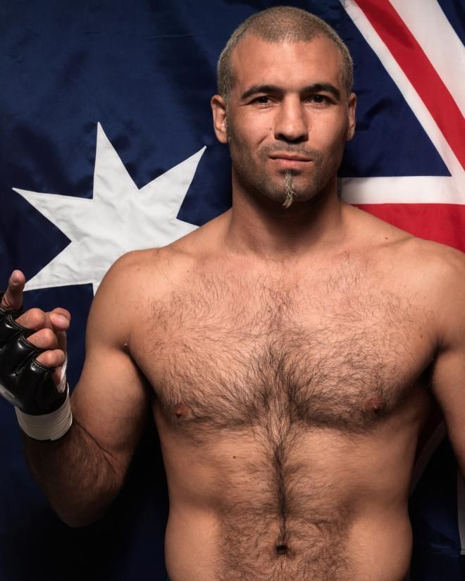 Man with a goatee poses shirtless, hand up, in front of an Australian flag.
