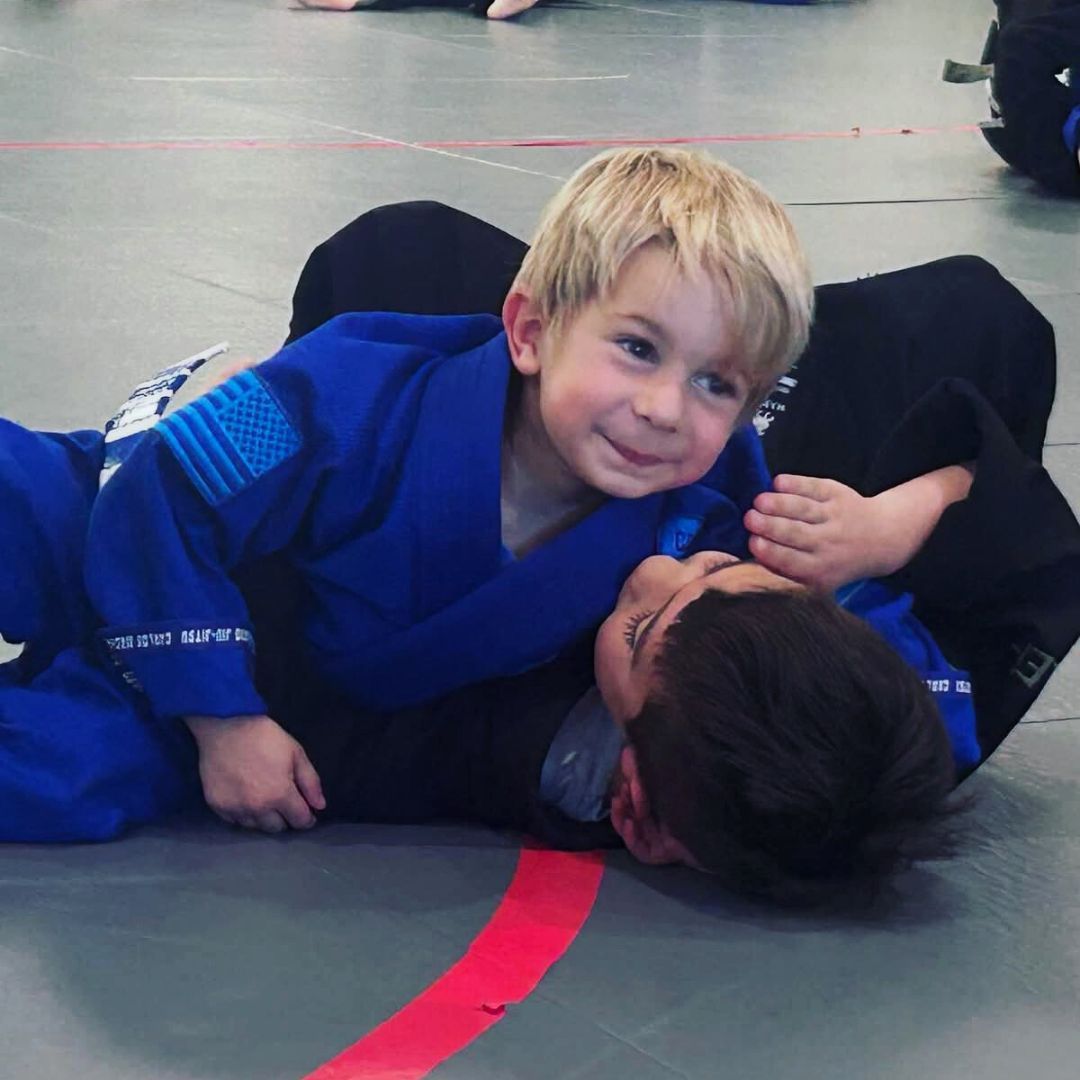 Two young children in blue and black martial arts uniforms grappling on a gym floor, one smiling.