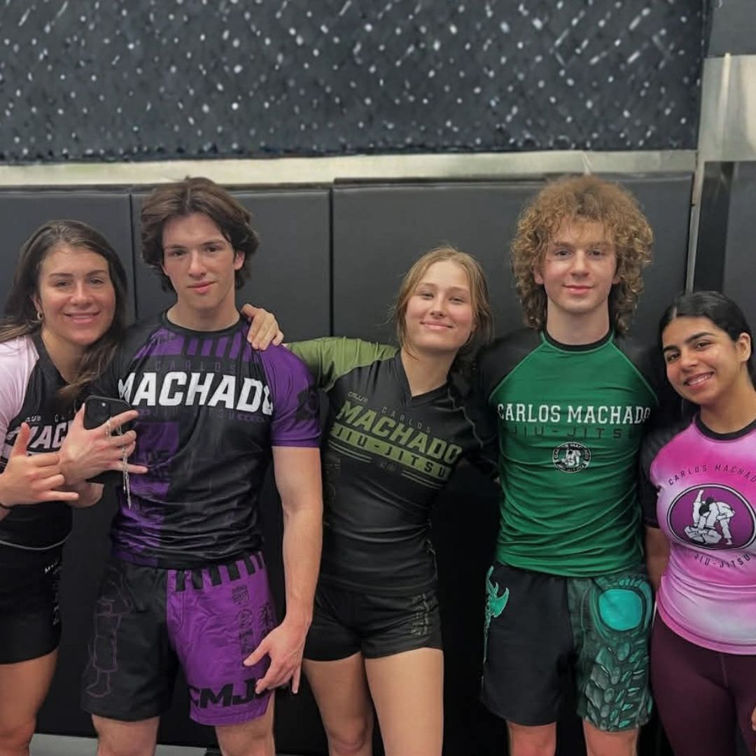 Five people in jiu-jitsu gear pose together in a gym.  Smiling,  some have arms around each other. Machado logo visible.