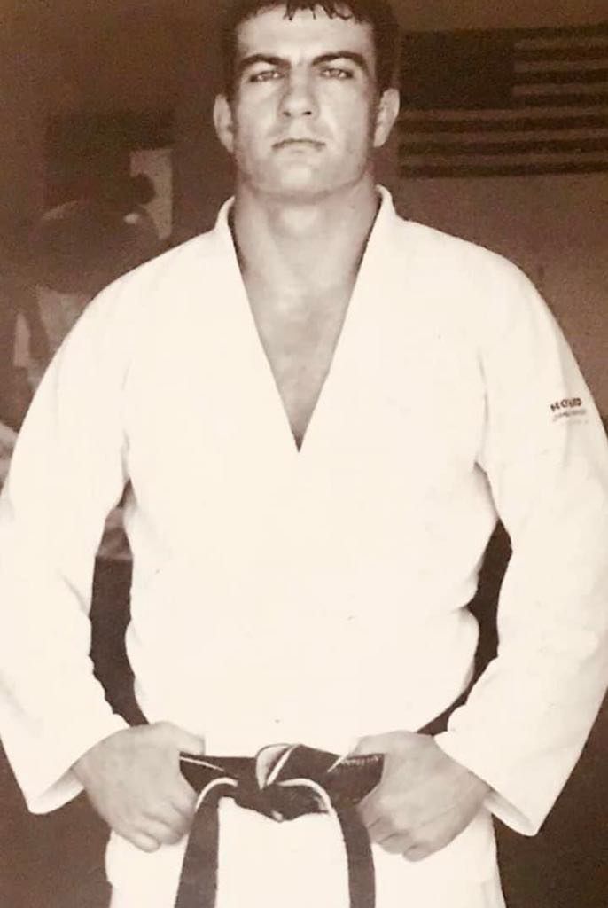 Man in judo gi, black belt, stands facing forward with hands on belt, American flag in background.