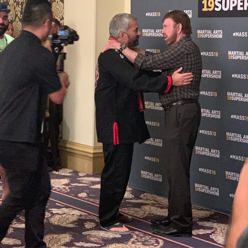 Chuck Norris hugs a man in a black martial arts uniform. A press area with a backdrop is visible.