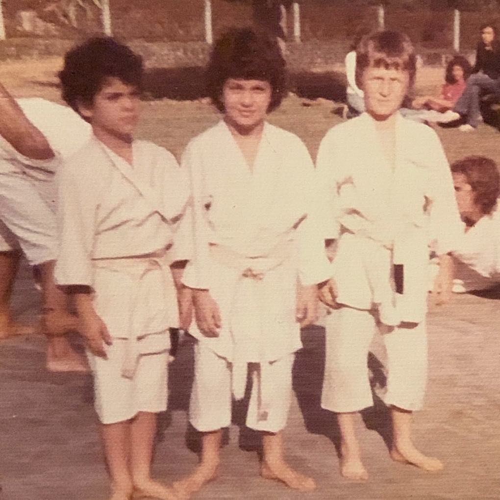 Three children in white martial arts uniforms stand outdoors, barefoot, with a blurred background.