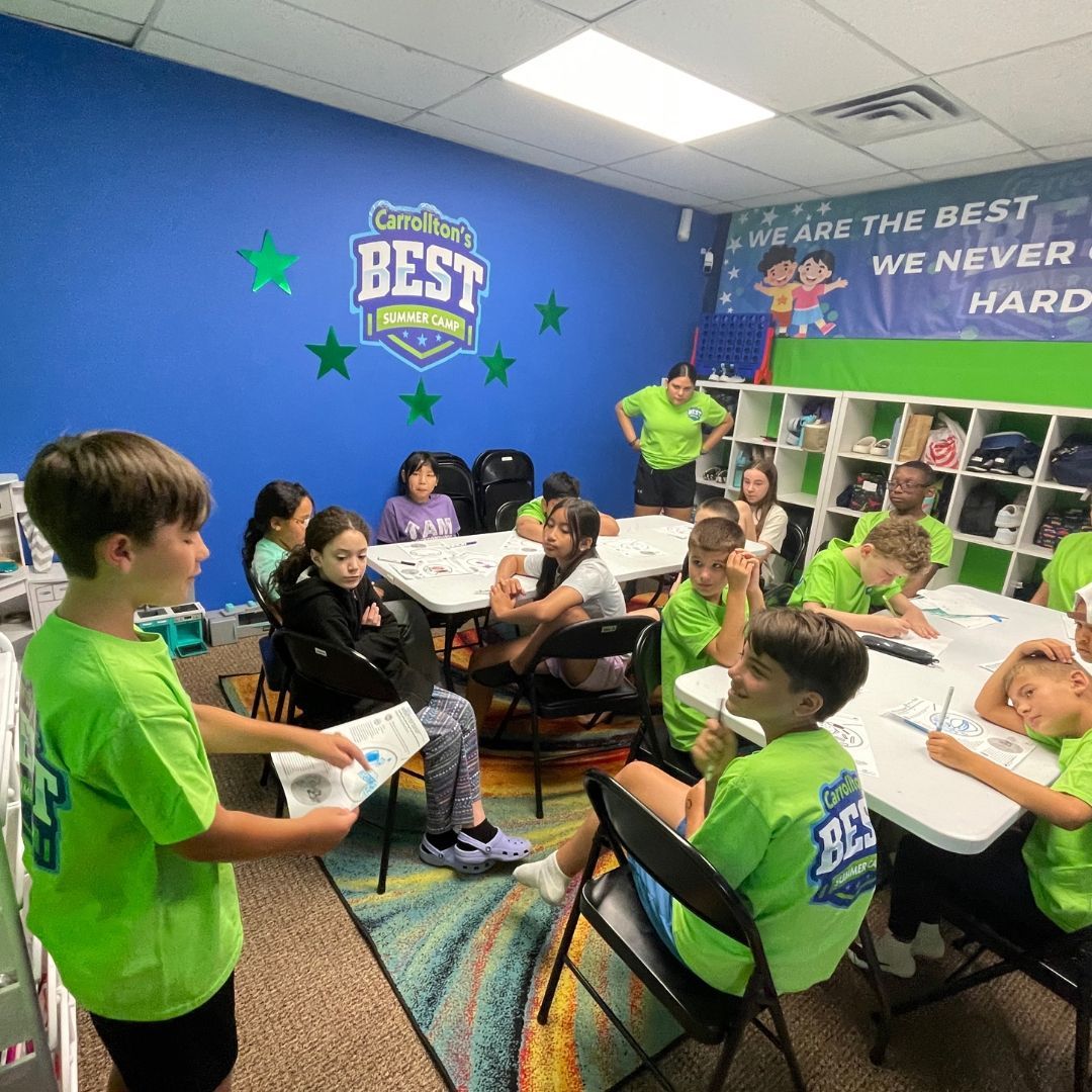Children in a classroom setting, some wearing green shirts, seated at tables. Blue walls with motivational banners.