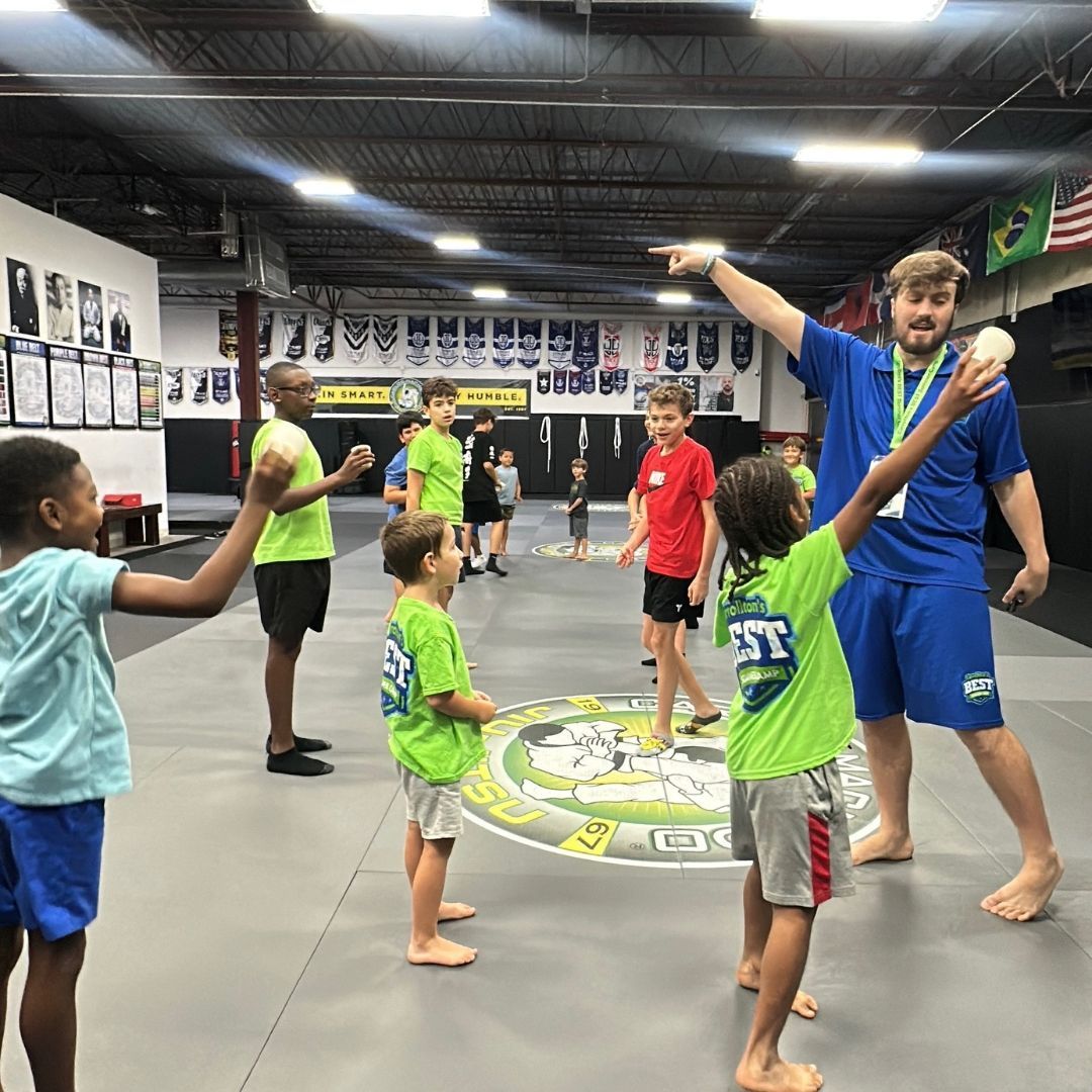 Children and instructor with balls inside a gym. Instructor points while children hold balls, wearing colorful shirts.