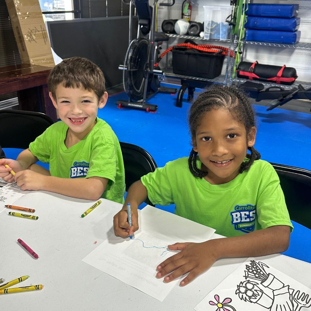 Two children smiling and coloring at a table; one boy and one girl. They are wearing green shirts, and the room has exercise equipment.