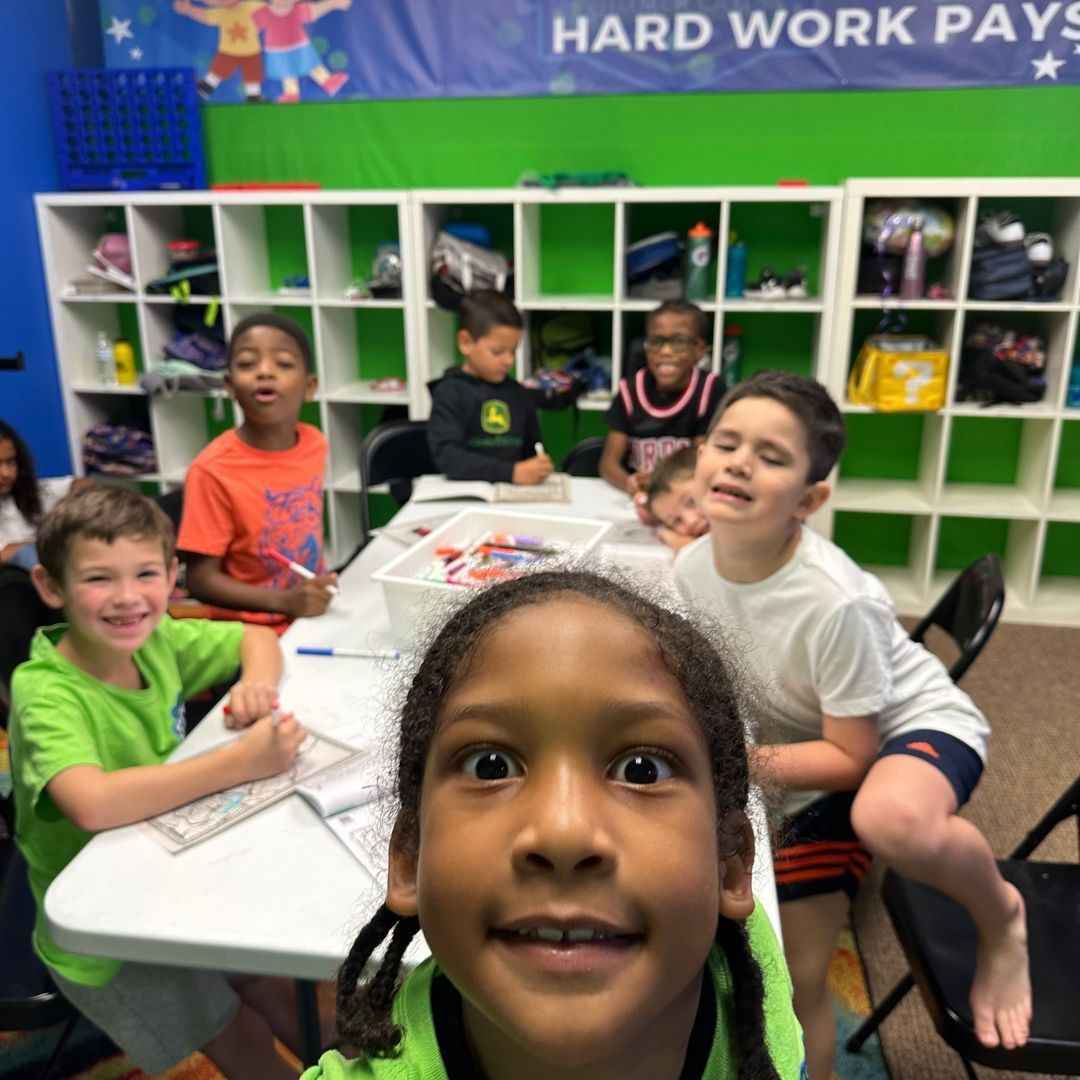 Group of kids smiling at the camera at a table in a classroom with white shelves.