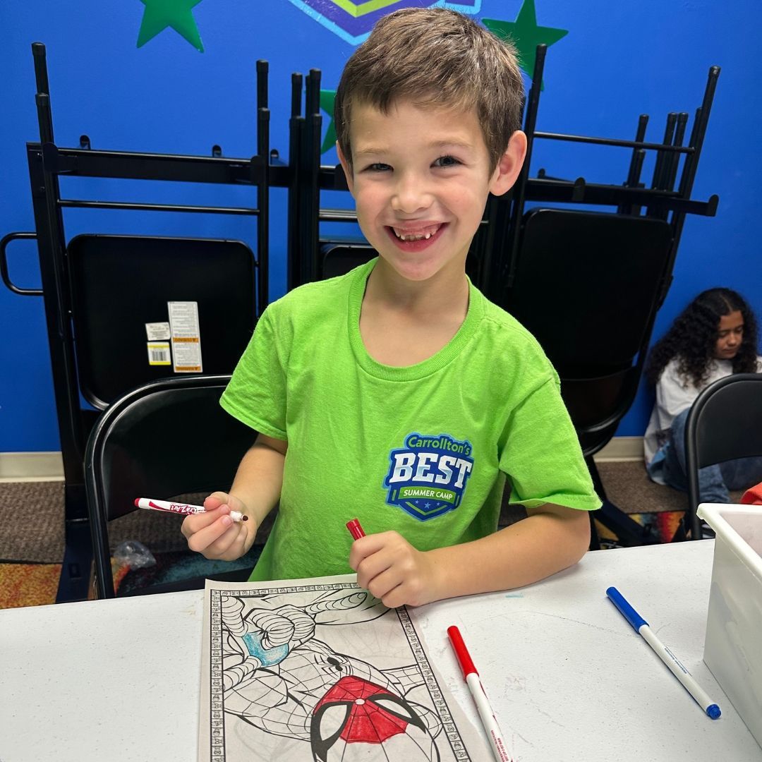 Boy smiling, coloring a Spider-Man picture with markers, at a table, in a room with a blue wall.