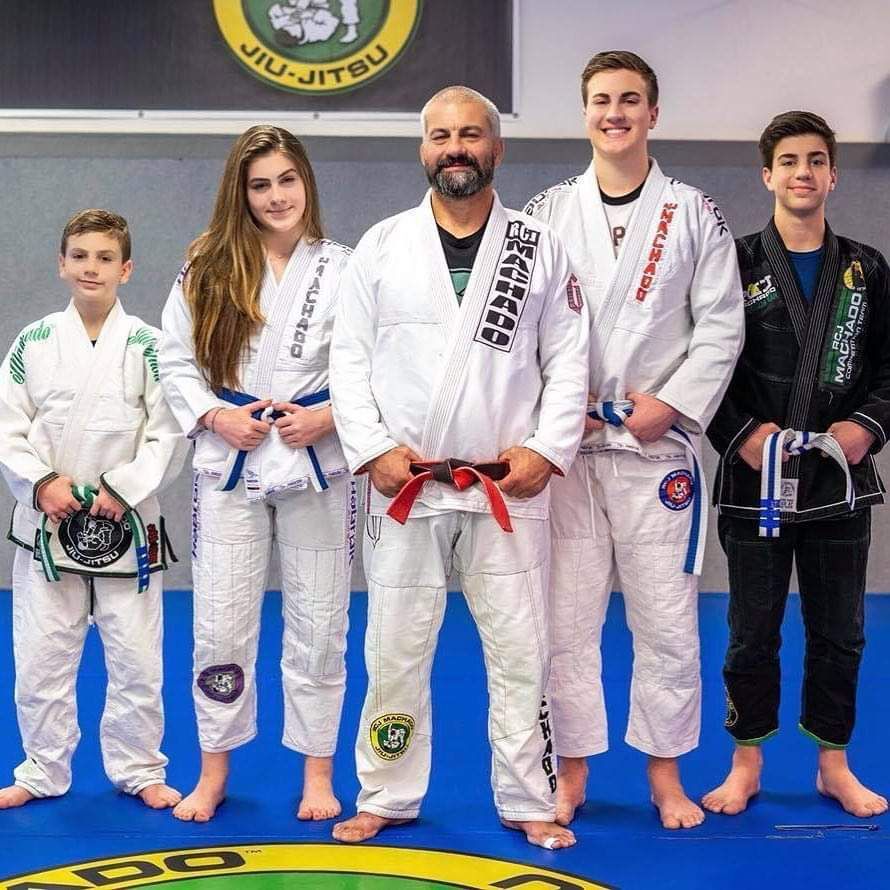 Five people in white and blue jiu-jitsu uniforms stand together on a blue mat. A man in the center wears a red belt.