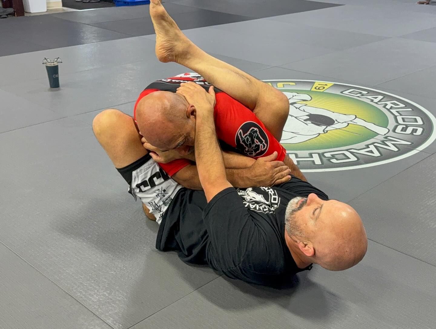 Two men in martial arts attire demonstrating a submission hold in a gym. One man is choking the other.