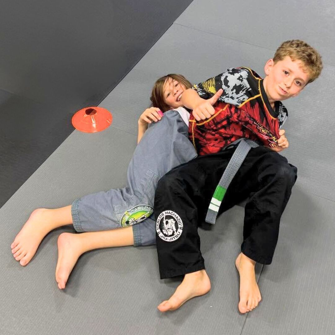 Two boys on a mat practicing Jiu-Jitsu. One boy is on top, giving a thumbs-up.