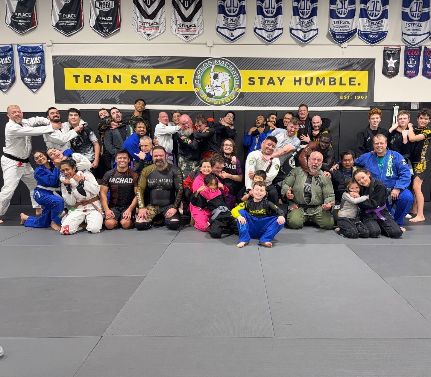Group of people in martial arts attire posing on a mat in a gym.