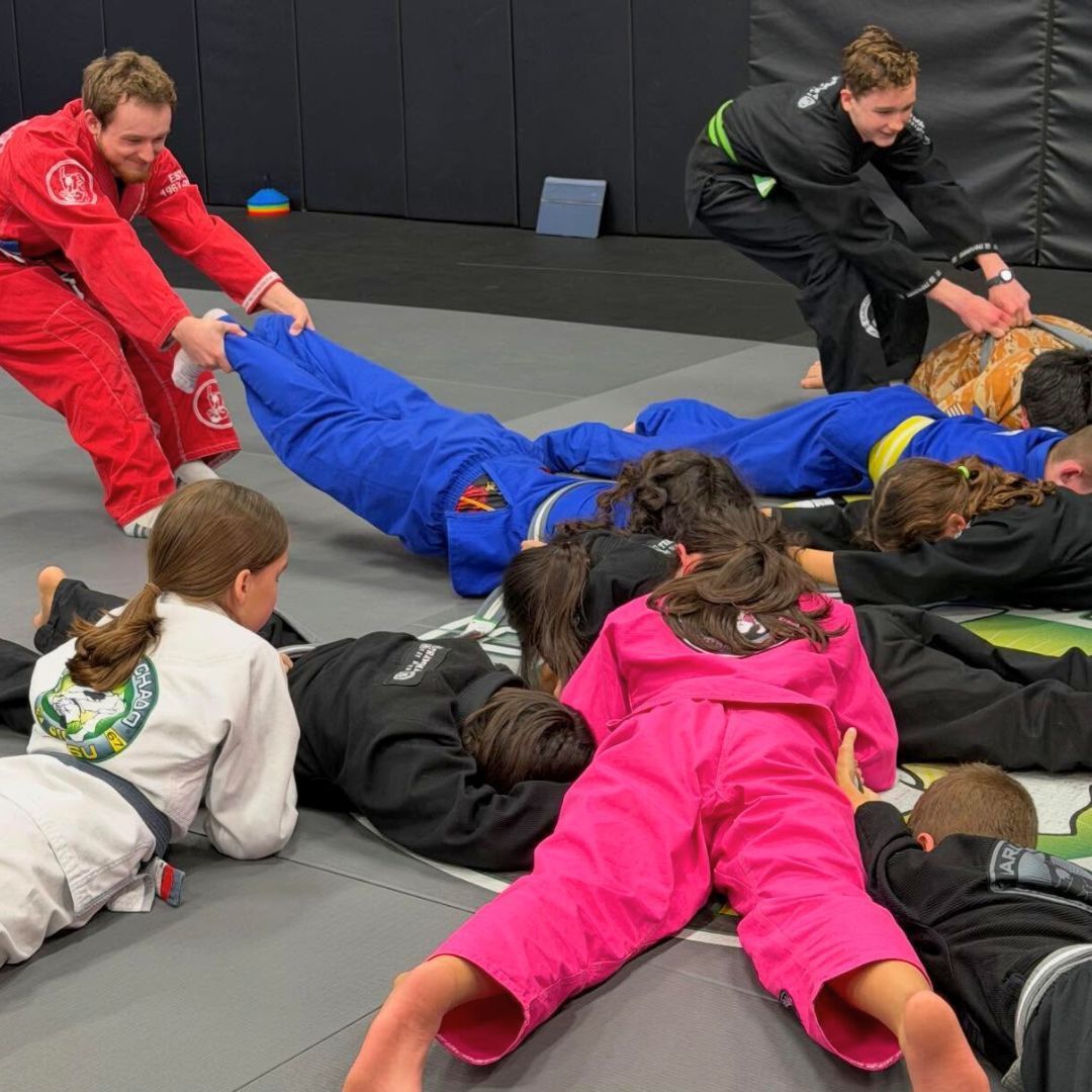 Children in colorful gis play a game on a mat. Two instructors pull a student in blue.