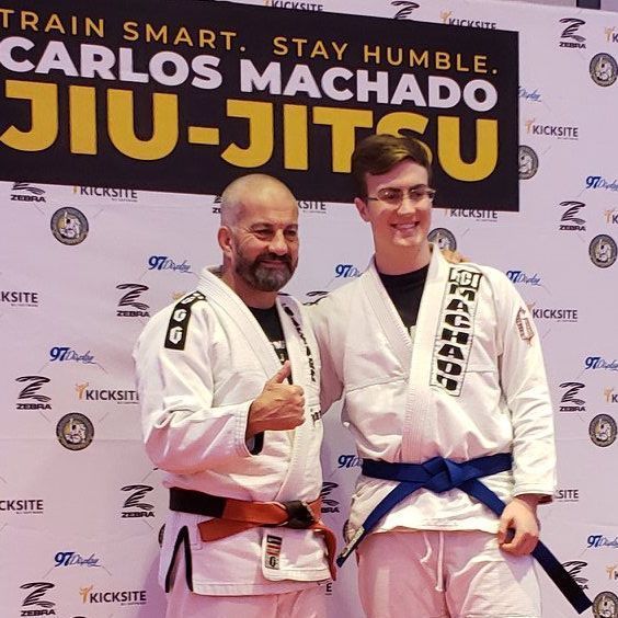Two men in white Jiu-Jitsu gis pose in front of a banner. One man gives a thumbs up.