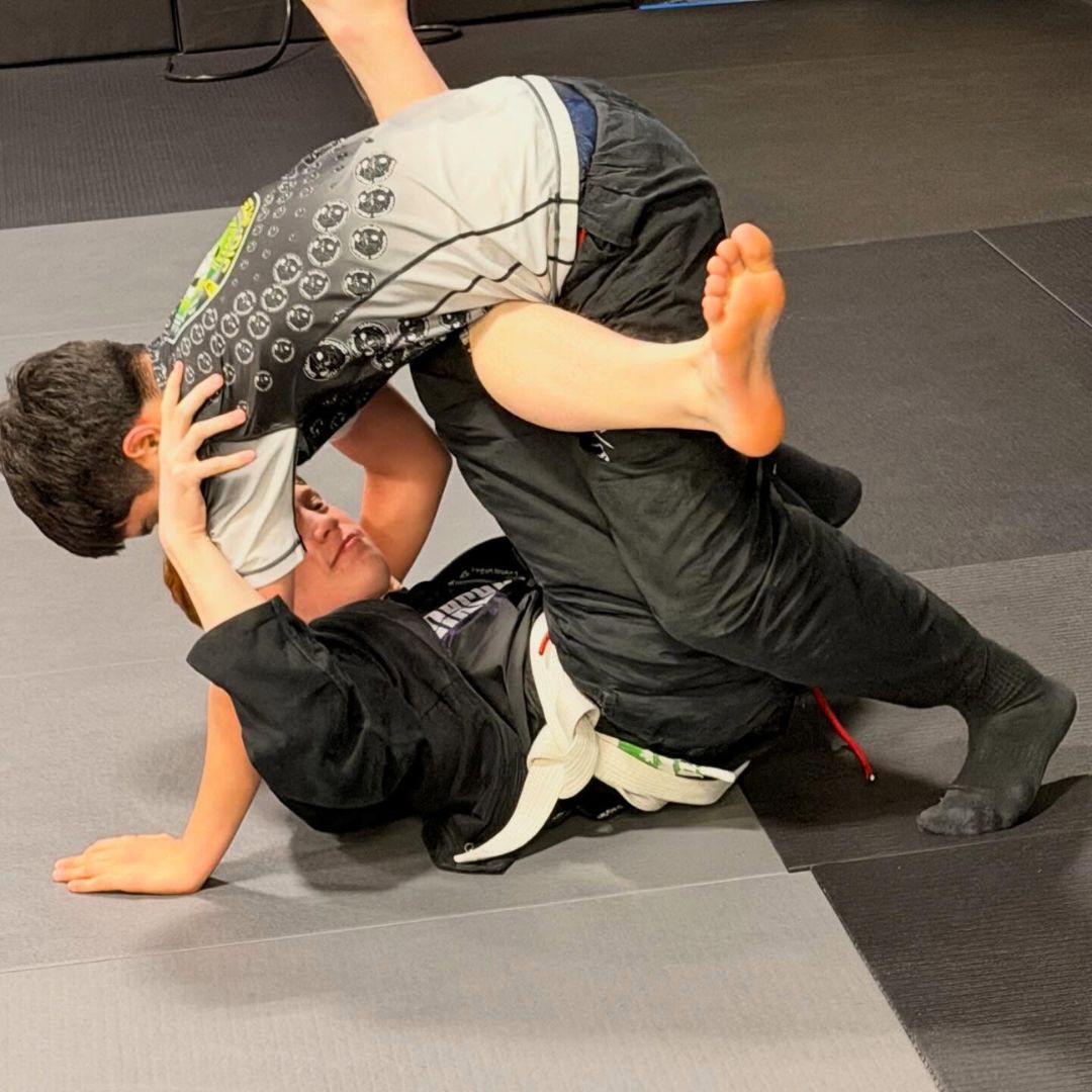 Two people grappling on a mat, one in a black gi, the other in a rash guard, legs entangled.