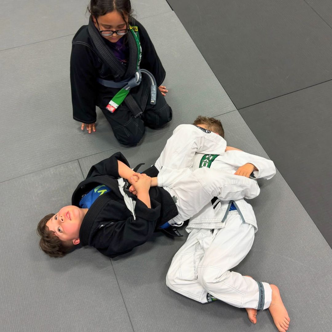 Two children practicing Brazilian Jiu-Jitsu on a mat. One child in white gi has the other in a leg lock. A girl watches.
