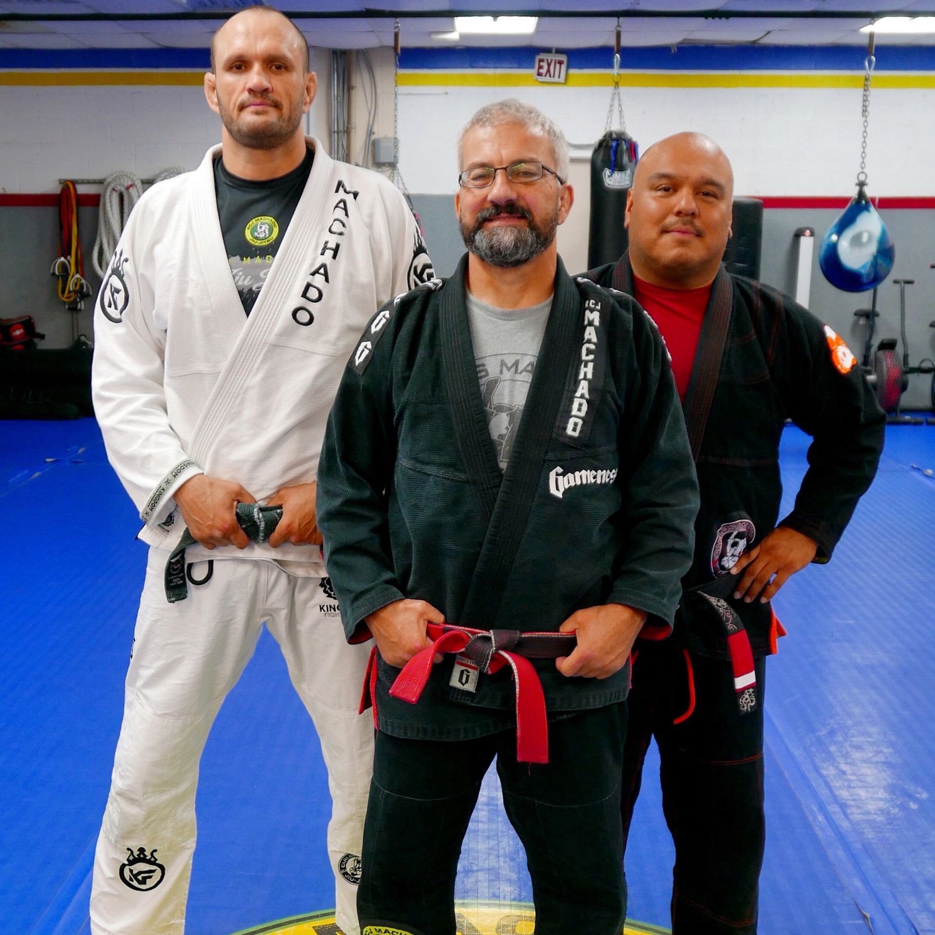 Three men in martial arts attire pose in a gym. The central man has a red belt.