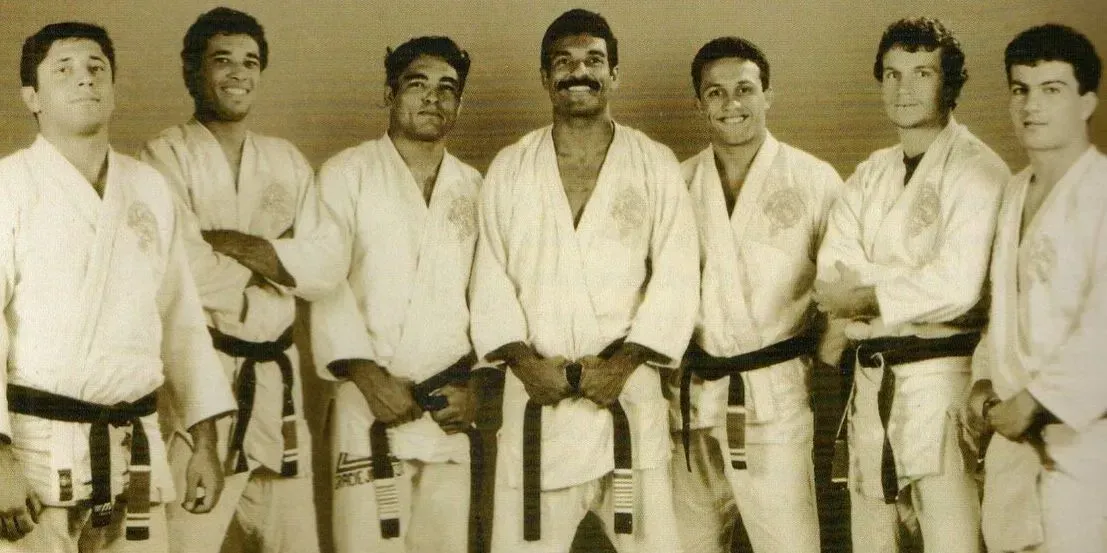 Group of seven people wearing white martial arts uniforms, black belts. They stand together, smiling.