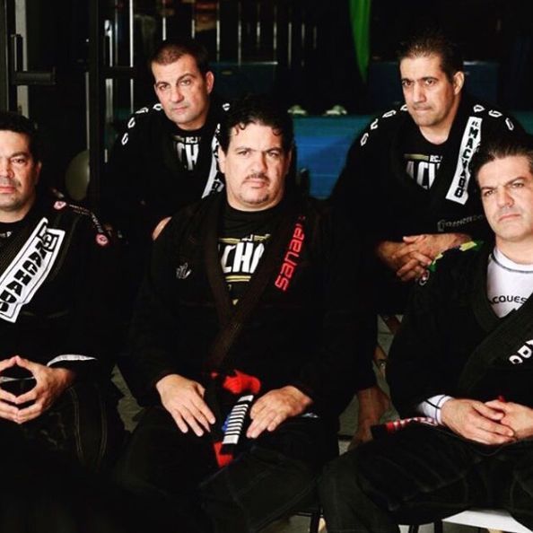 Group of five people in black martial arts uniforms, some with black belts, in a gym setting.