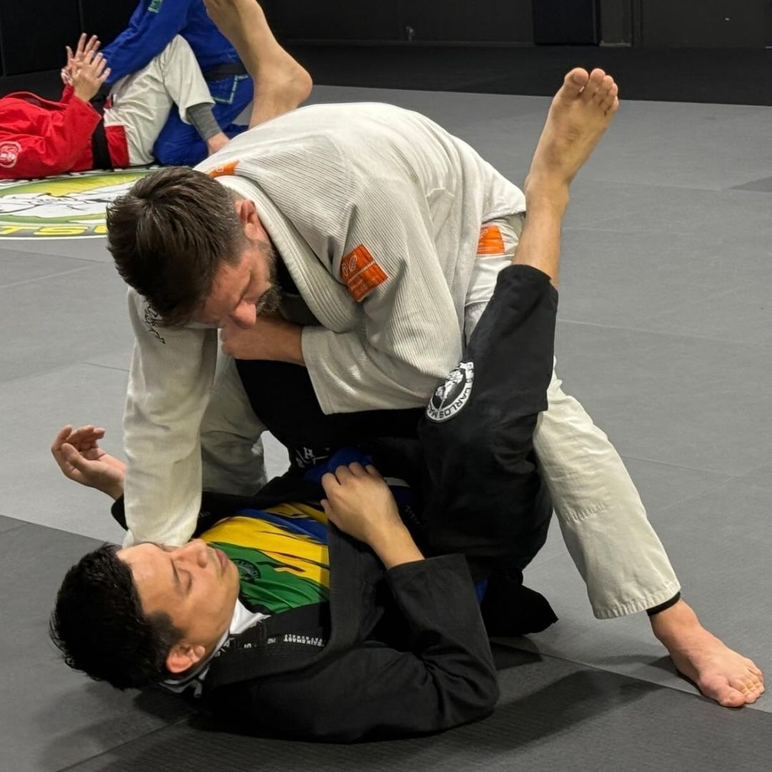 Two men grappling on a mat, one in a white gi, the other in a black gi, inside a training gym.