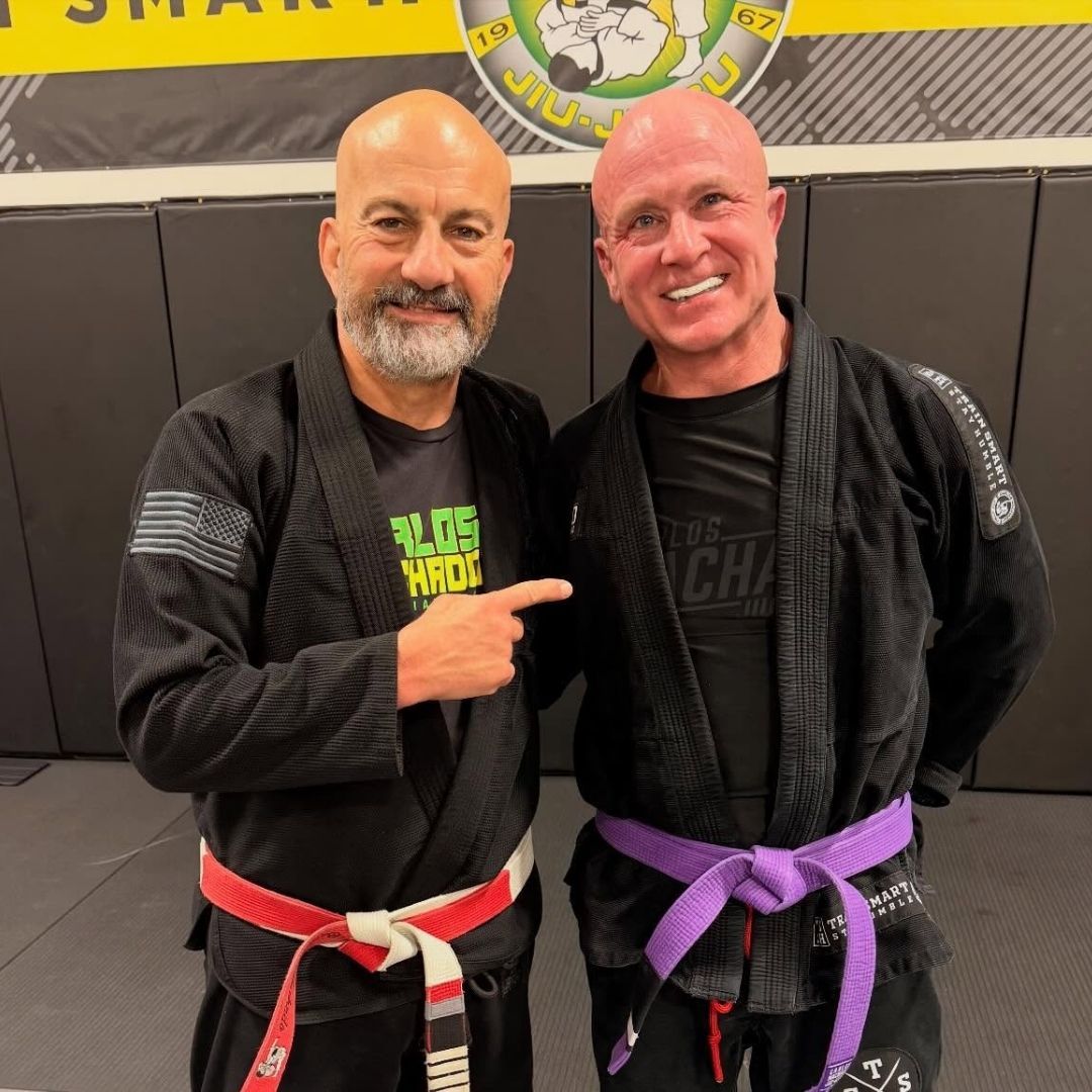 Two men in martial arts uniforms, one pointing at the other. Indoor setting with mats. Red and purple belts.