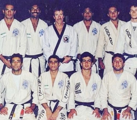 Group of men in martial arts uniforms, some black belts. Chuck Norris in the center, white gi with black belt.