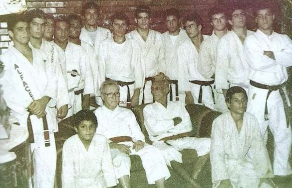 Group of karate practitioners in white uniforms, posing with two seated men, outdoors.