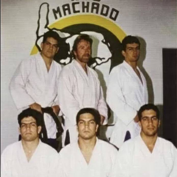 Chuck Norris with six people wearing white martial arts uniforms in front of a MACHADO logo.