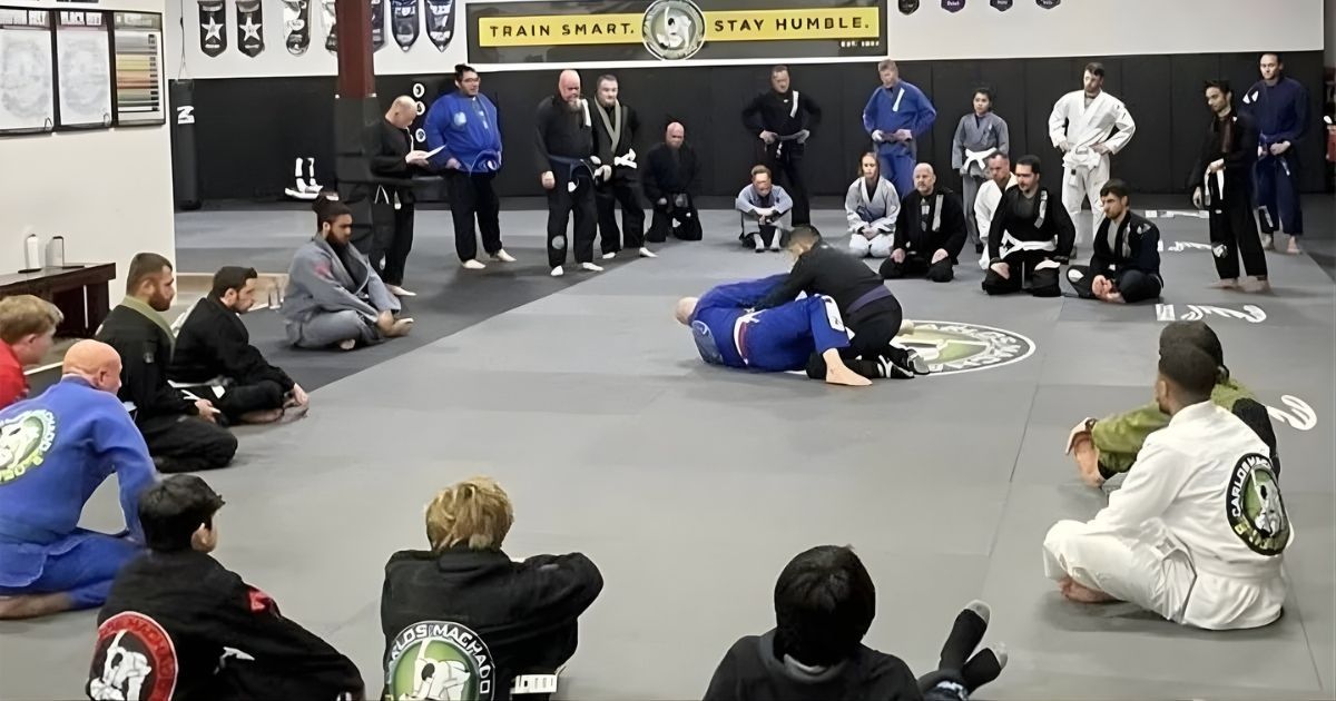 People in martial arts gi uniforms observing a grappling demonstration on a mat in a gym.