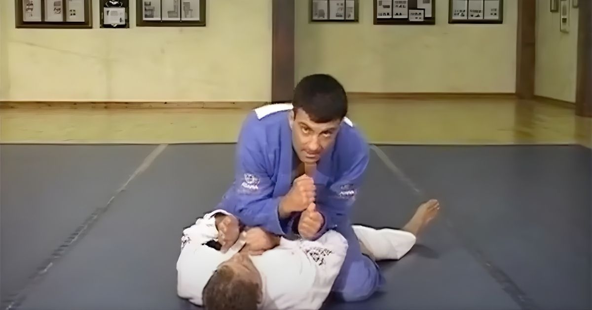 Man in blue gi demonstrating a Jiu-Jitsu move on a man in a white gi, indoors on a mat.