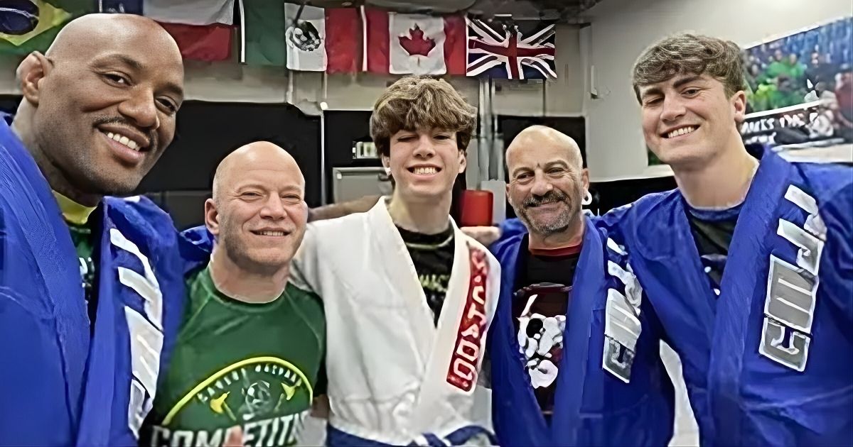 Five people smiling, one in a white gi, others in blue gis, standing in front of flags.