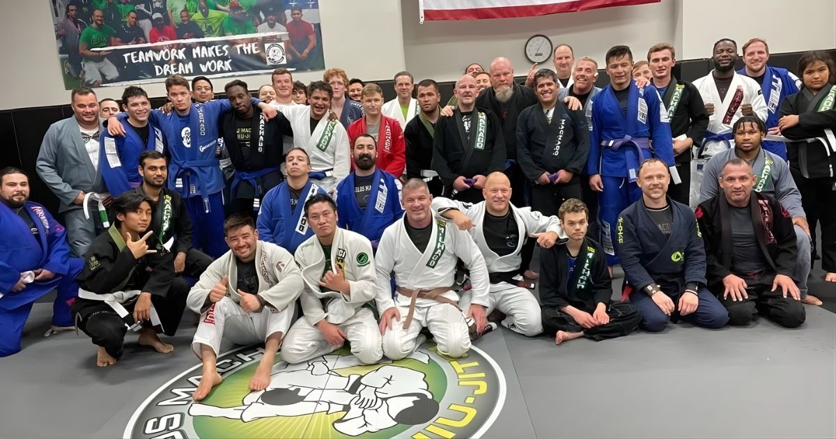Group of people in martial arts attire posing on a mat, indoors. Many wearing blue and white gis.