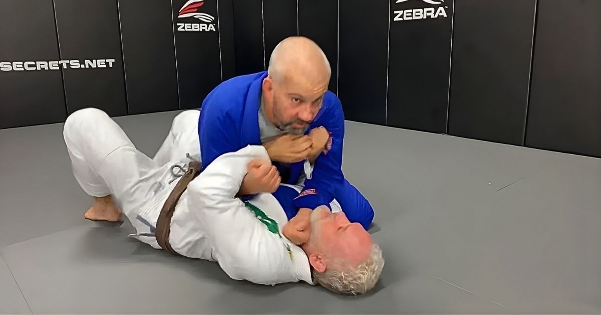 Two men in martial arts uniforms practice a grappling technique on a mat. One is on the ground.