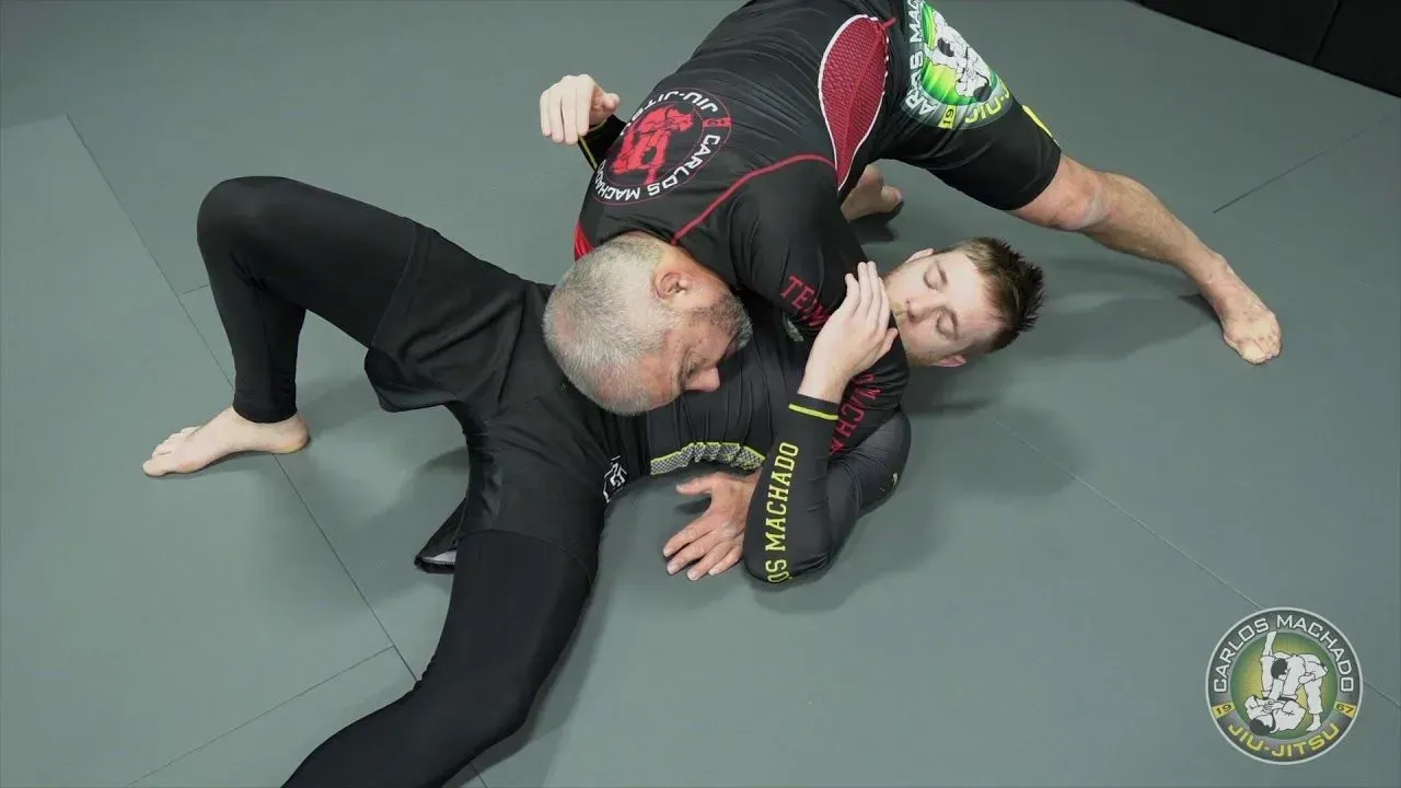 Two men grappling on a gray mat. One is in a dominant position, applying a choke.
