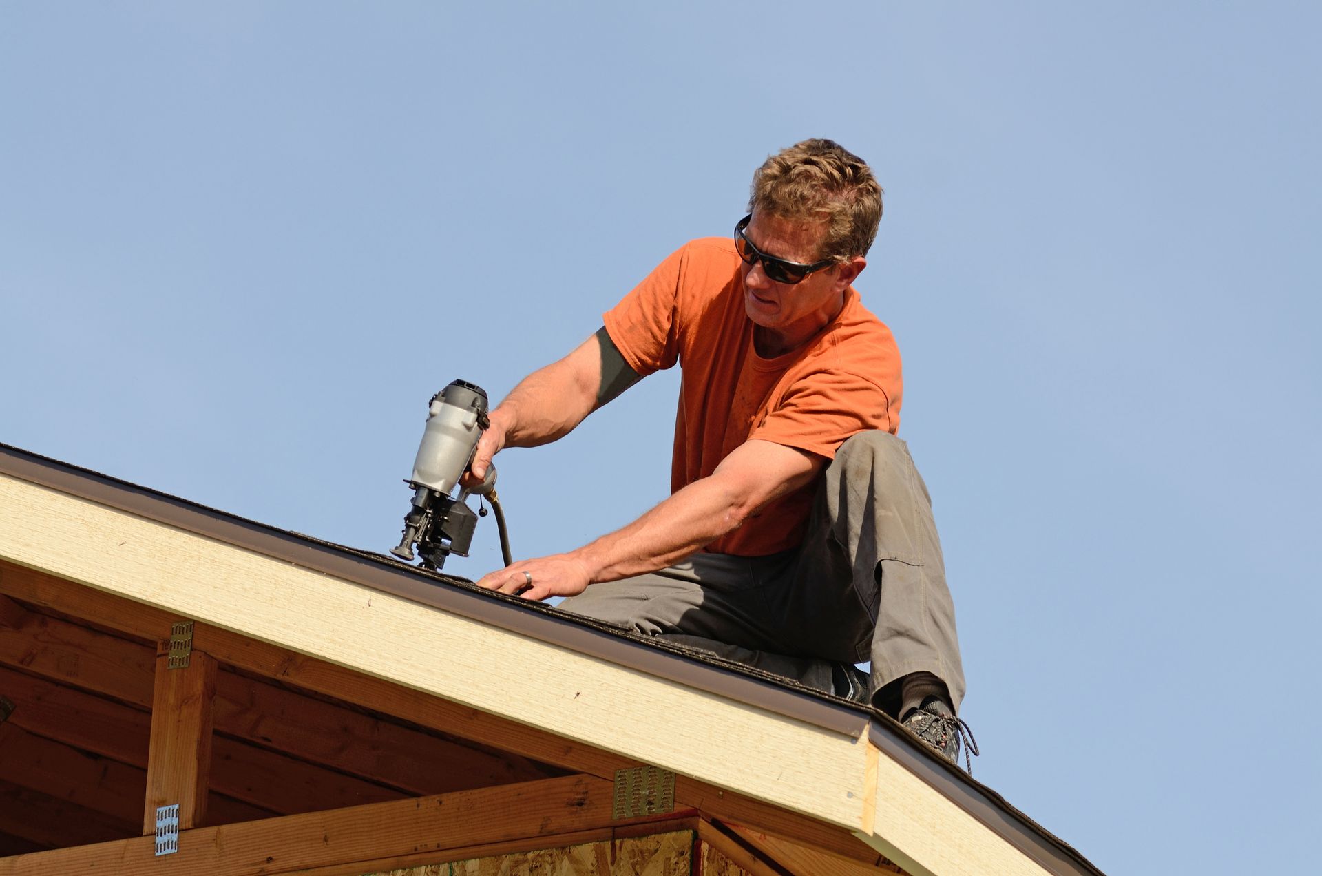 A mid-aged contractor wearing orange t-shirt and sunglasses is completing a roofing repair. A mid-aged contractor wearing orange t-shirt and sunglasses is completing a roofing repair.