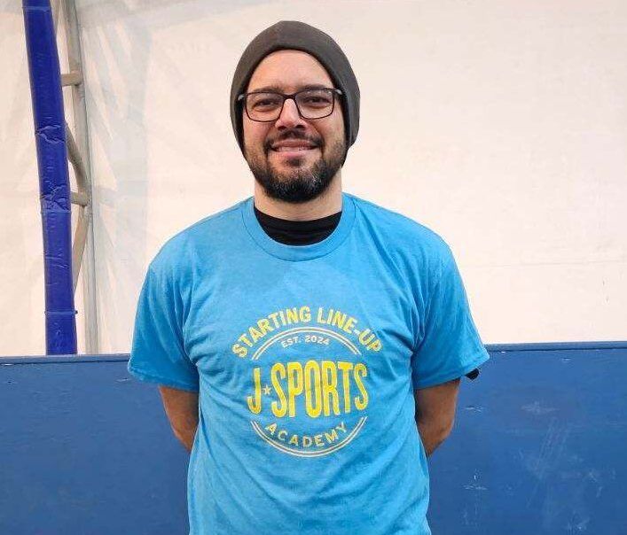 A man with a beard and glasses is wearing a blue shirt that says starting line up j-sports academy.