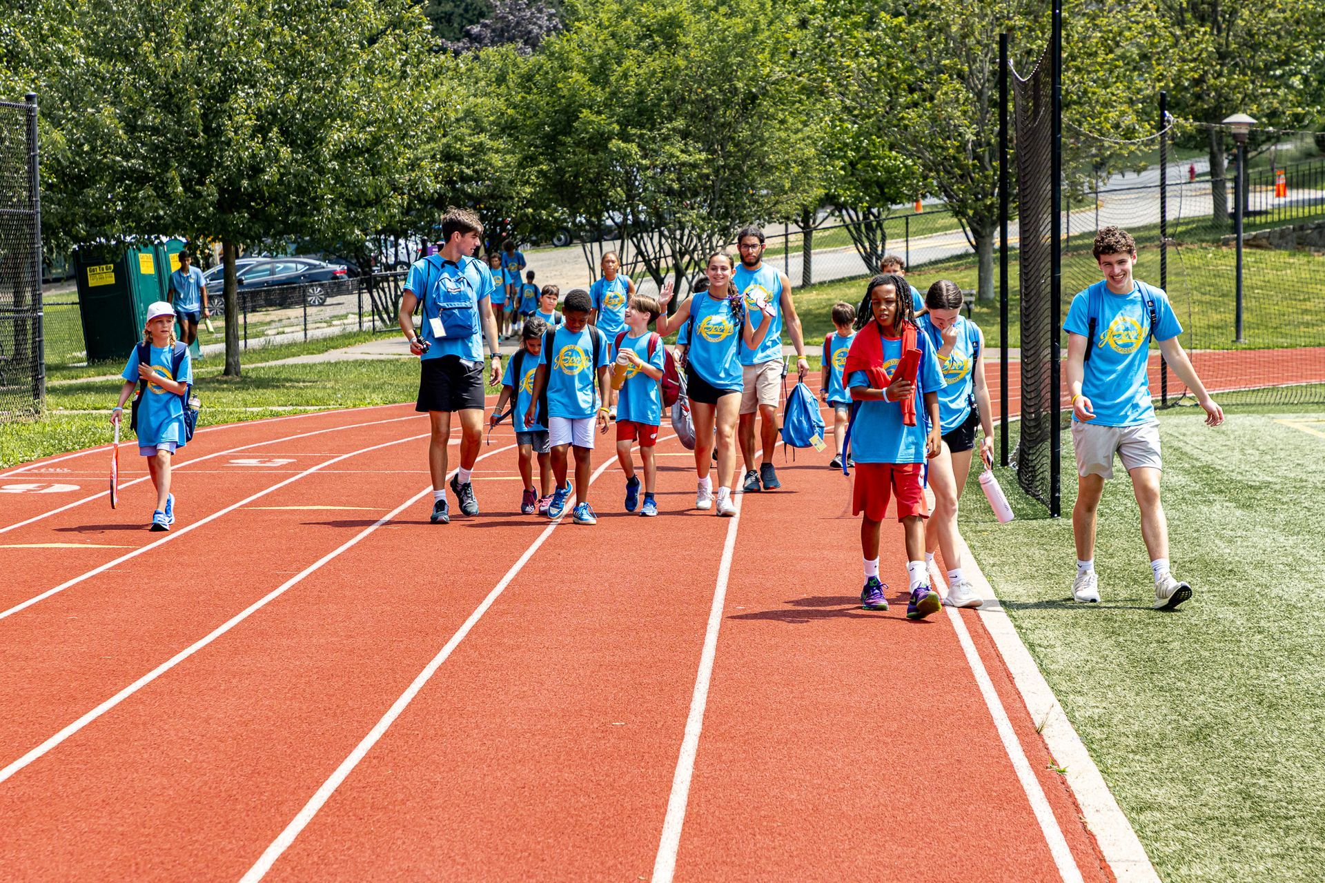 A group of children and adults in blue shirts walking on a red track in a sunny outdoor setting.