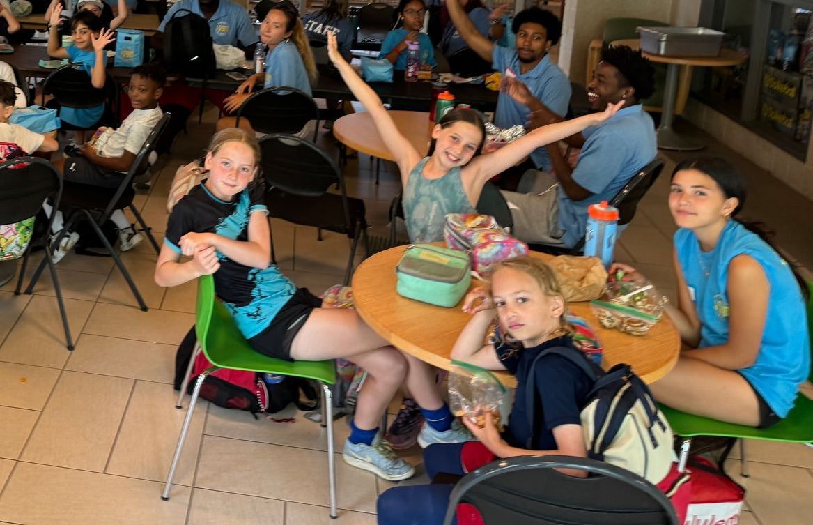 A group of children are sitting around a table with their arms in the air.