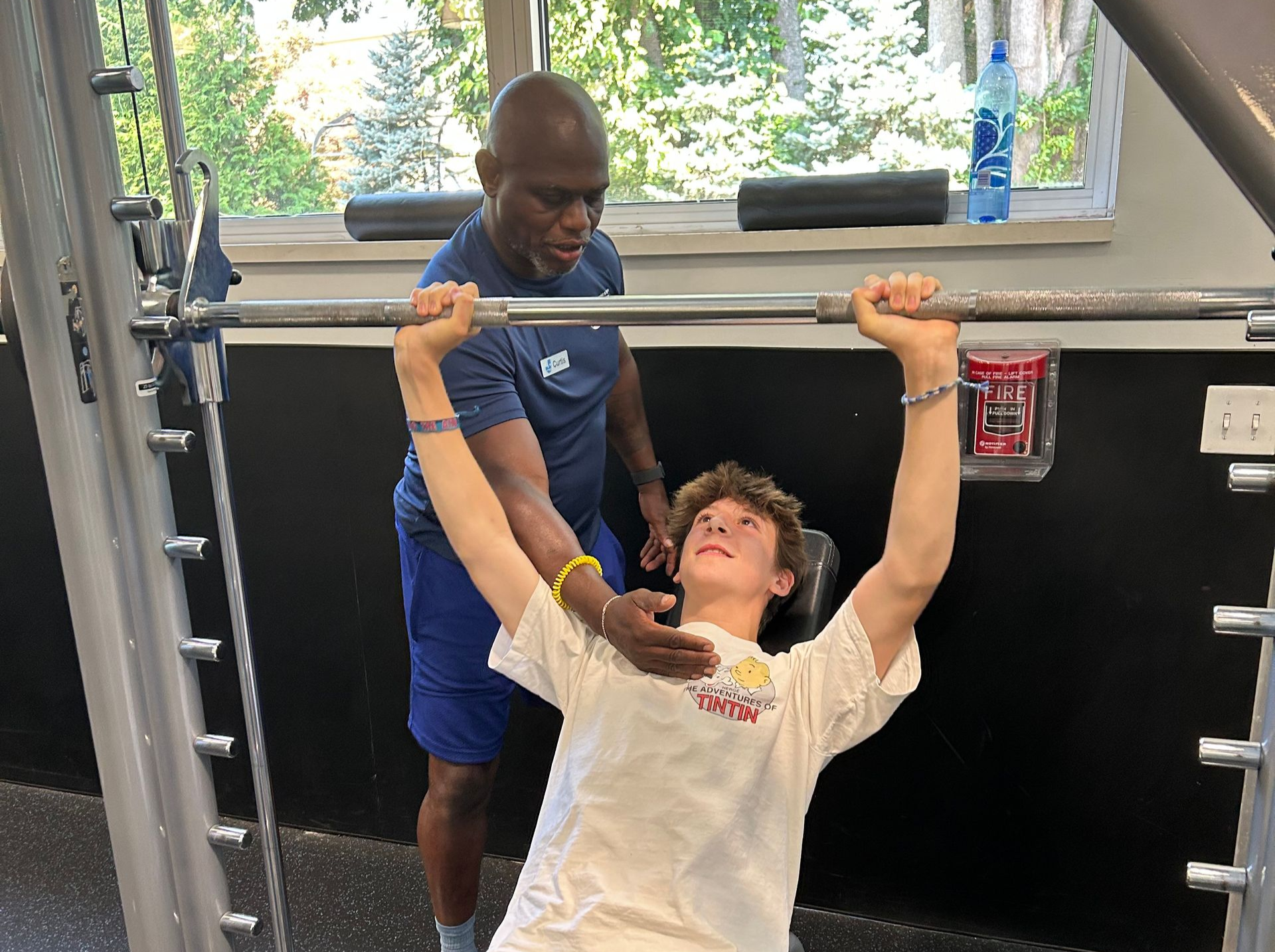 A man spots a young person bench pressing in a gym. Both have focused expressions.
