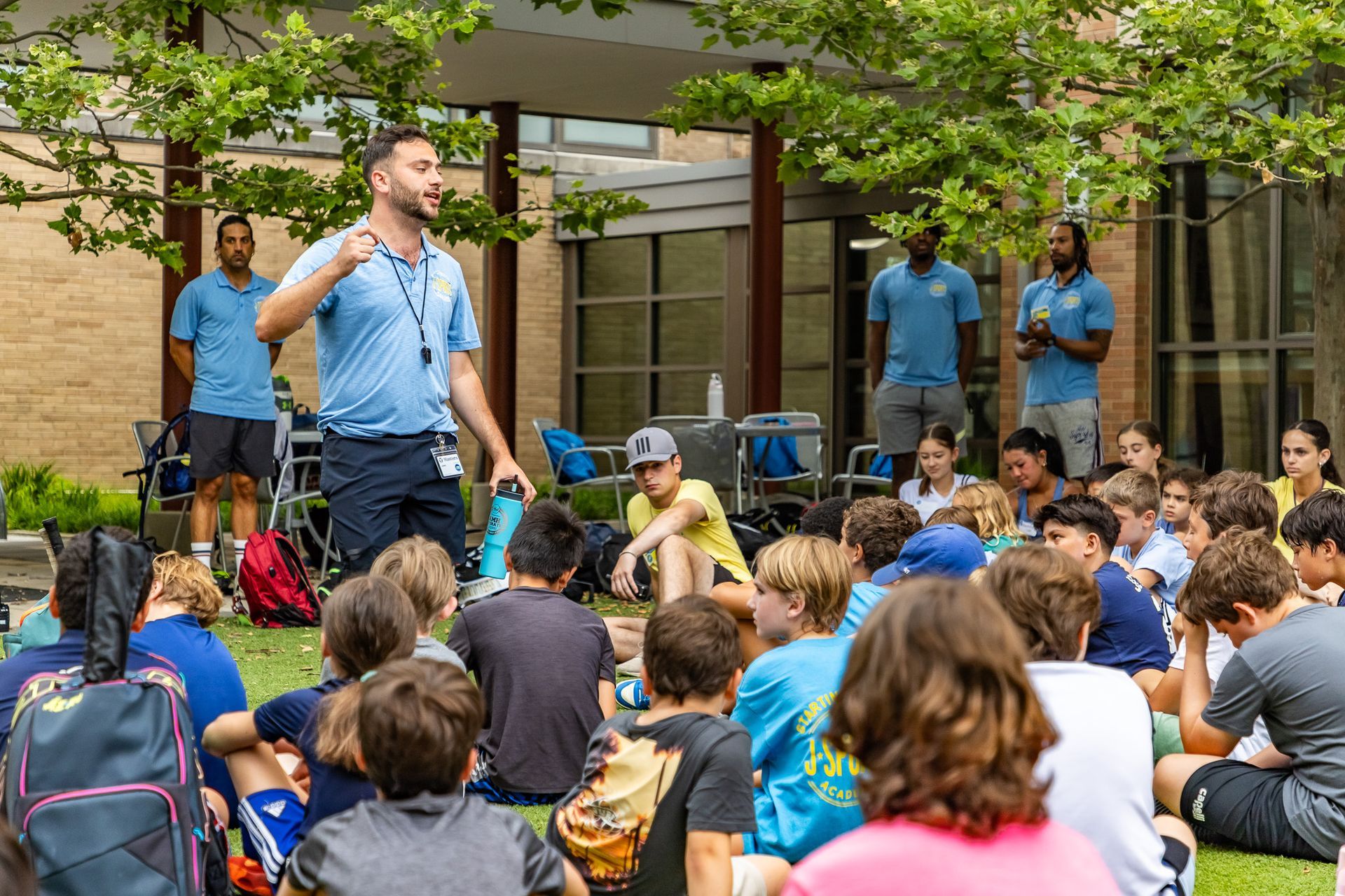 A man speaks to a group of children sitting on grass outside. Two other men stand behind them.