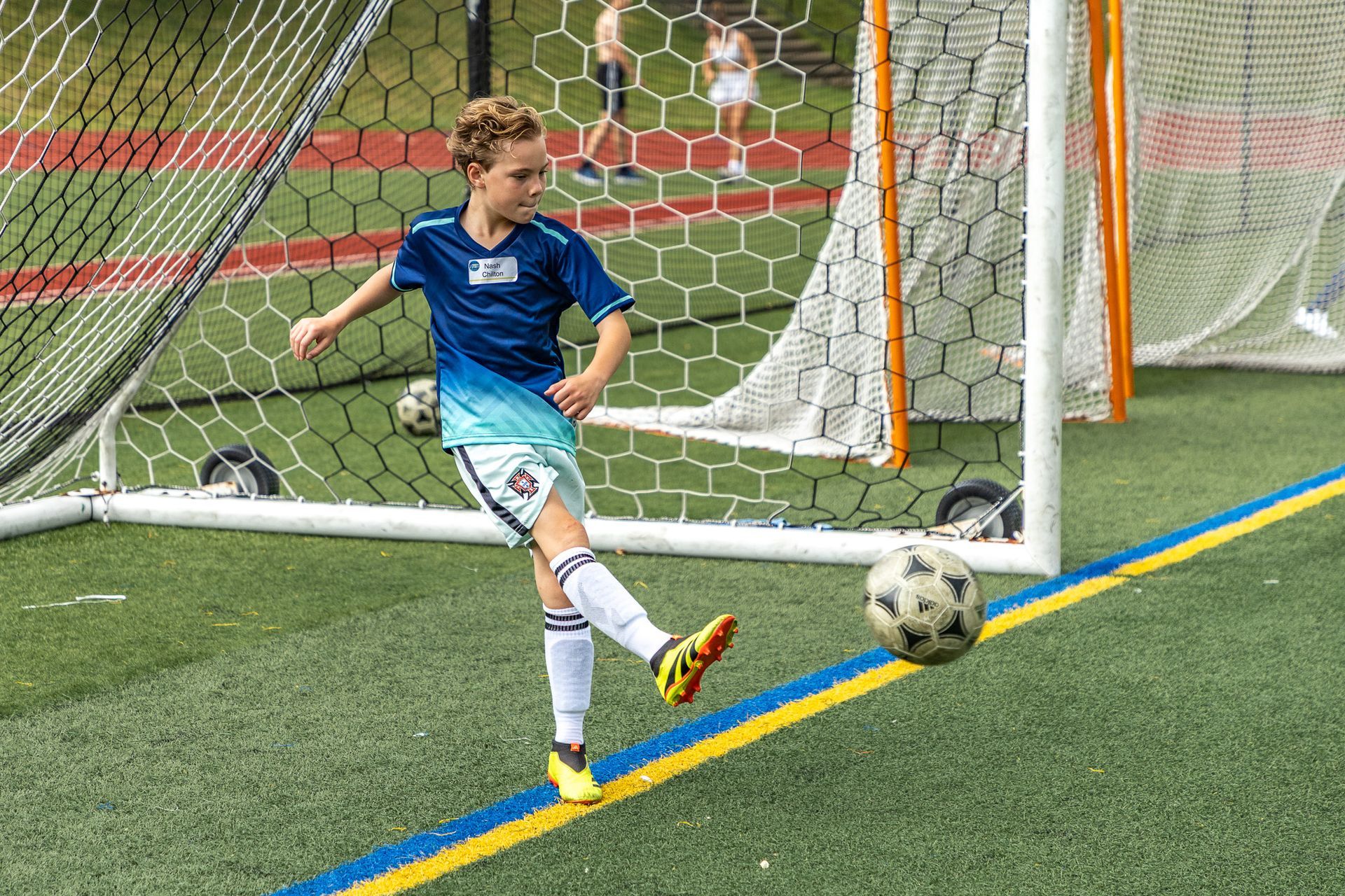 Boy in blue soccer uniform kicking a ball toward a goal on a green field. Other players and spectators are in the background.