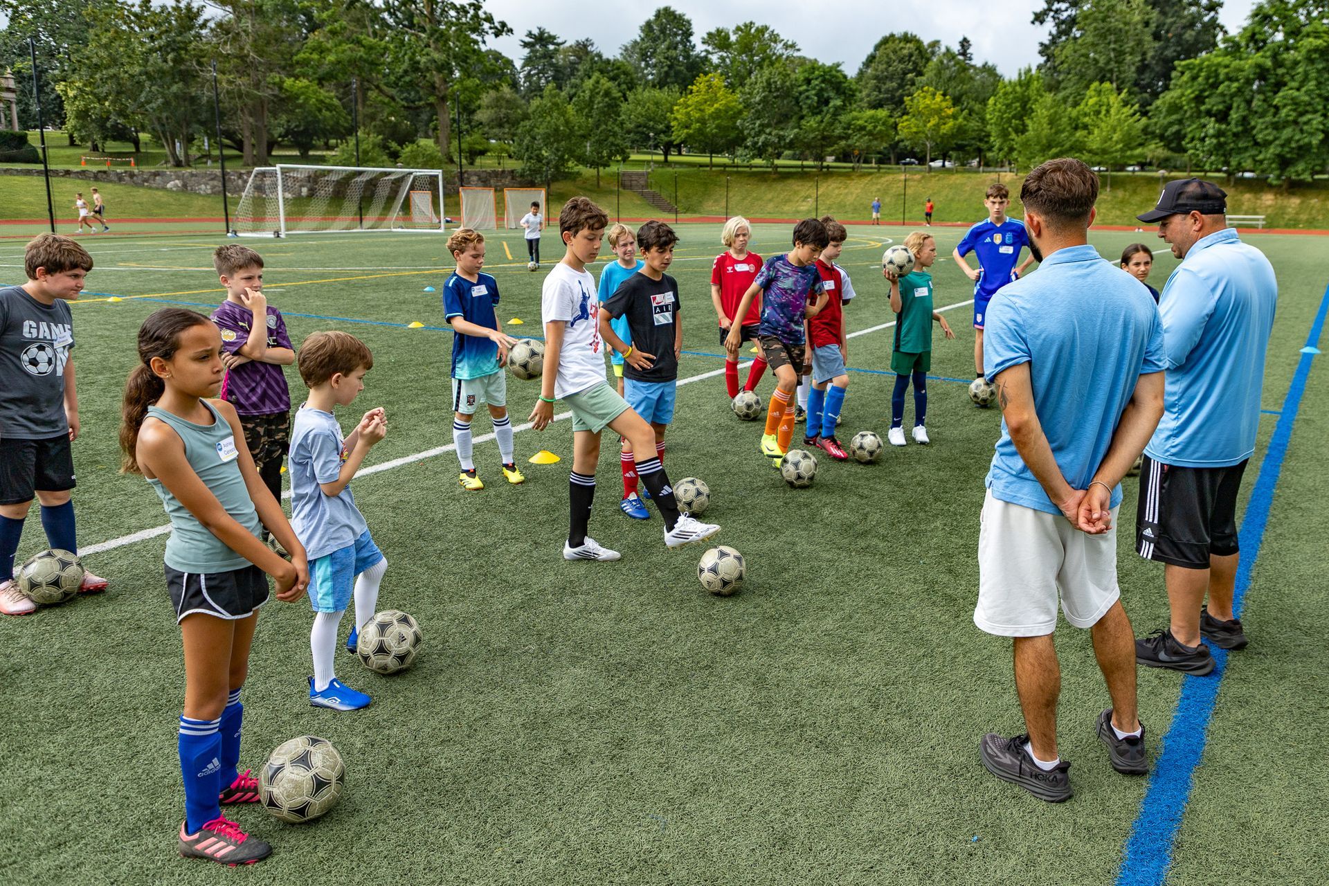A soccer coach instructs a group of children on a green turf field. Players are positioned with soccer balls.