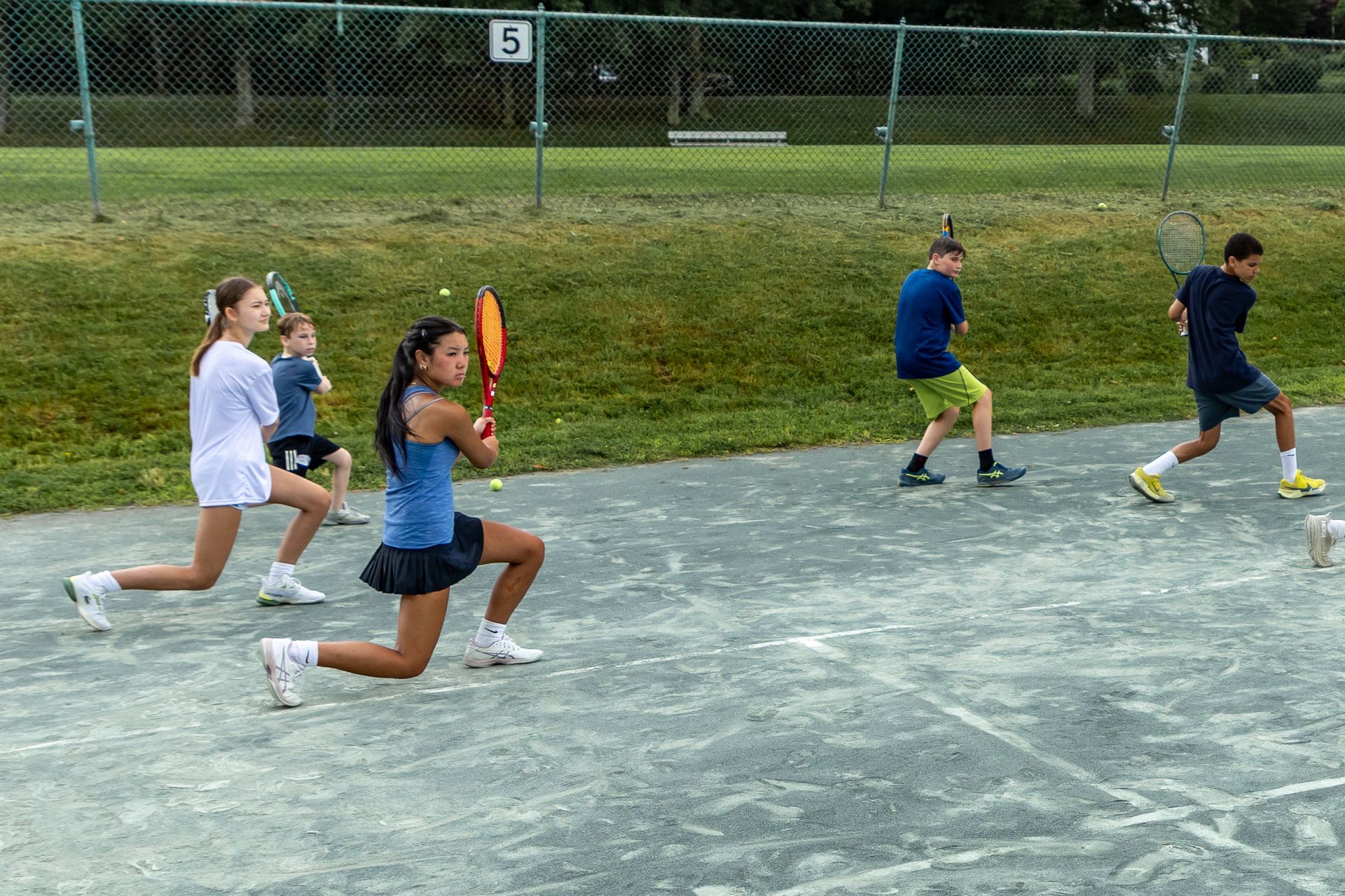 Tennis players practicing, on a court, some lunging. Green grass and fence in the background.