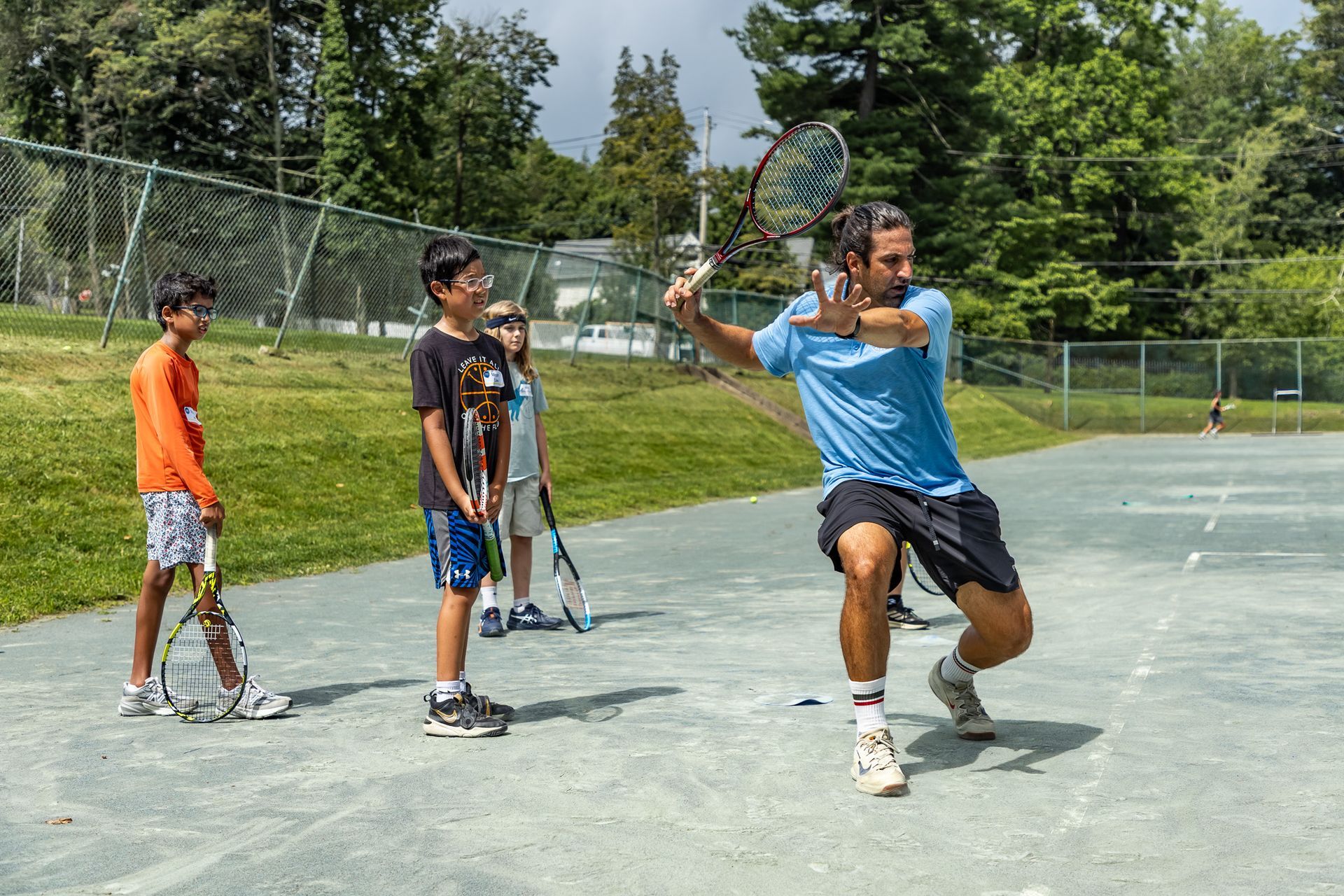 Tennis coach demonstrating a forehand swing to three young students on a court. Green grass, blue sky.