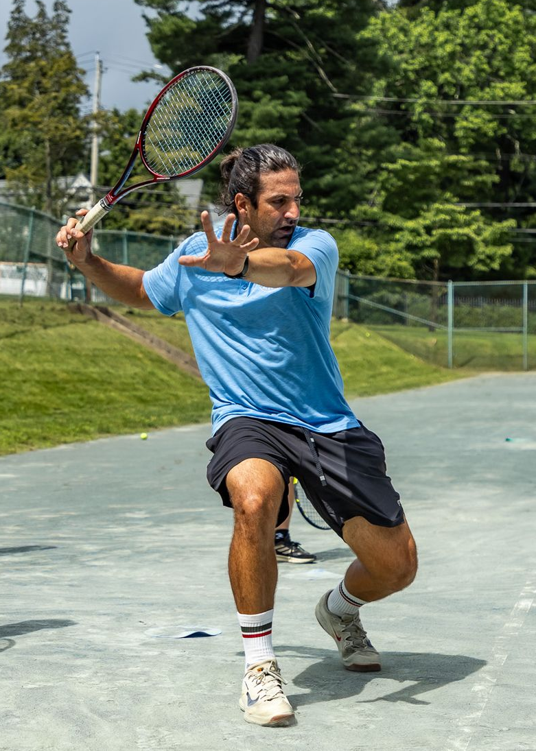 Man in blue shirt playing tennis, hitting a forehand on an outdoor court.