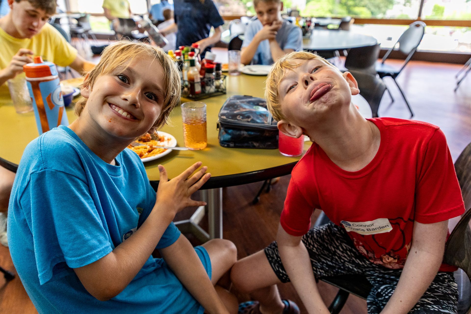 Two boys at a table, one smiling in blue, the other with food on his face in red. They are in a cafeteria.
