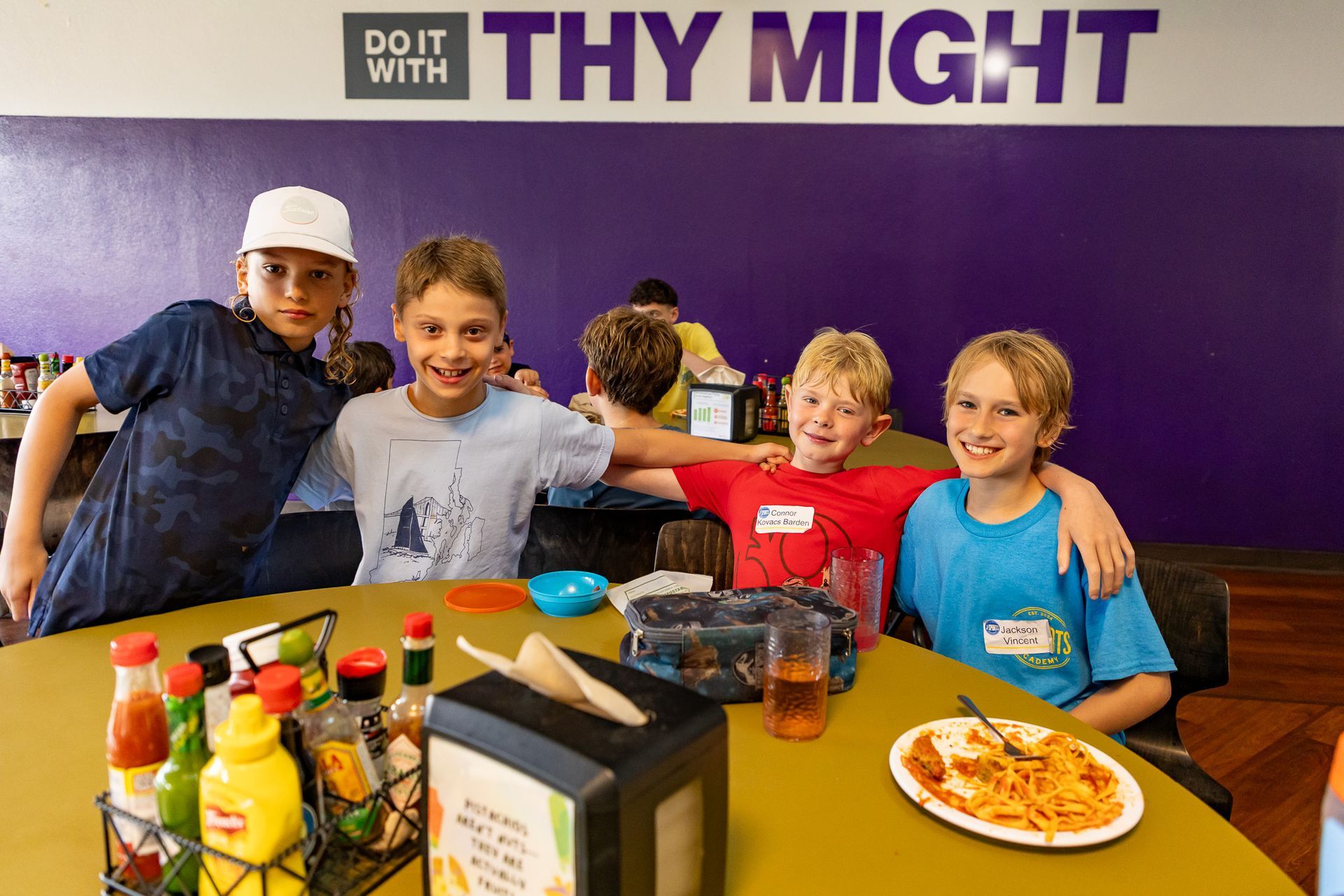 Four boys smiling at a table in a cafe, with food and condiments. Purple wall with “Do it with Thy Might” written on it.