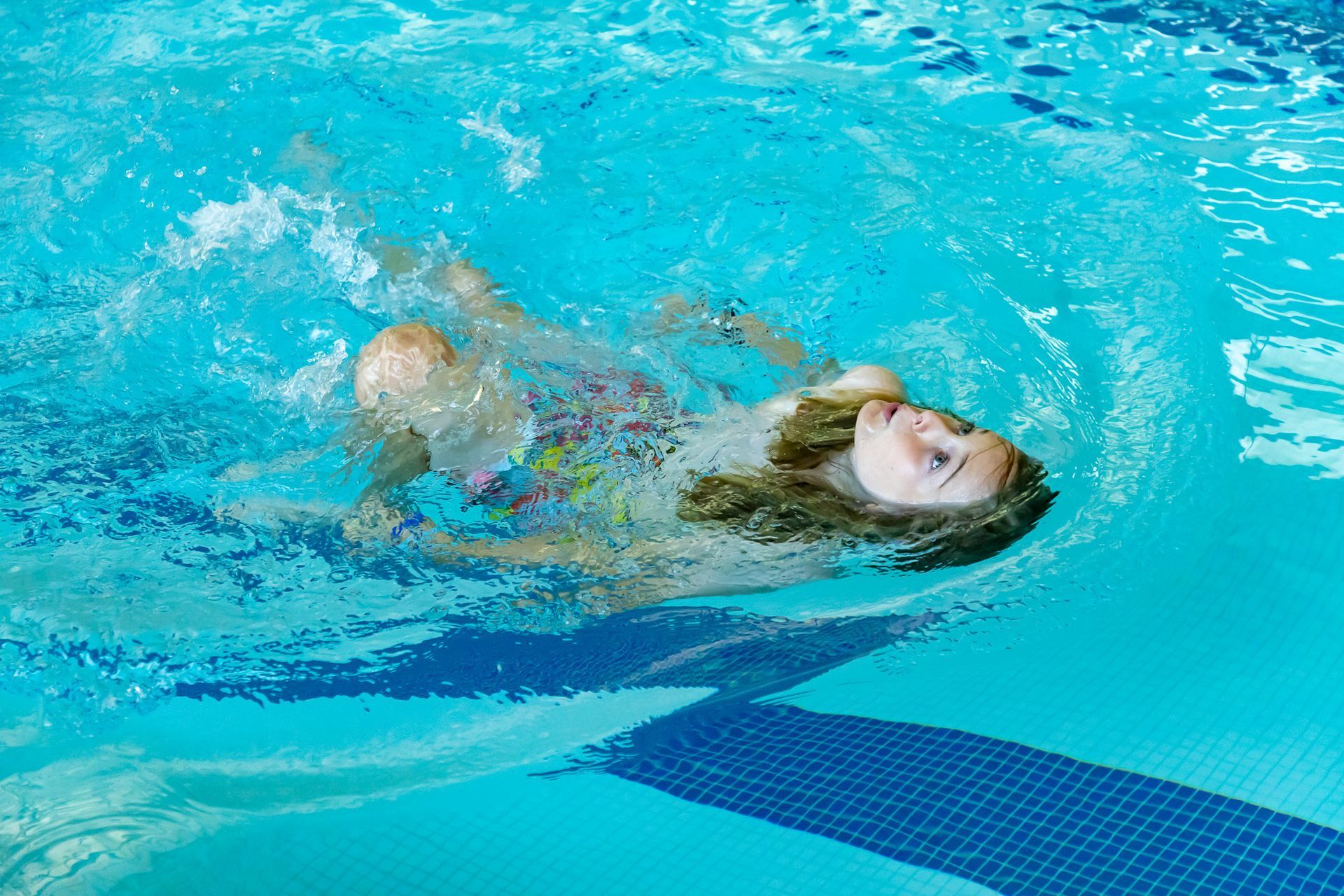 Girl in a pool splashing, her face above the water; blue water, sunny setting.