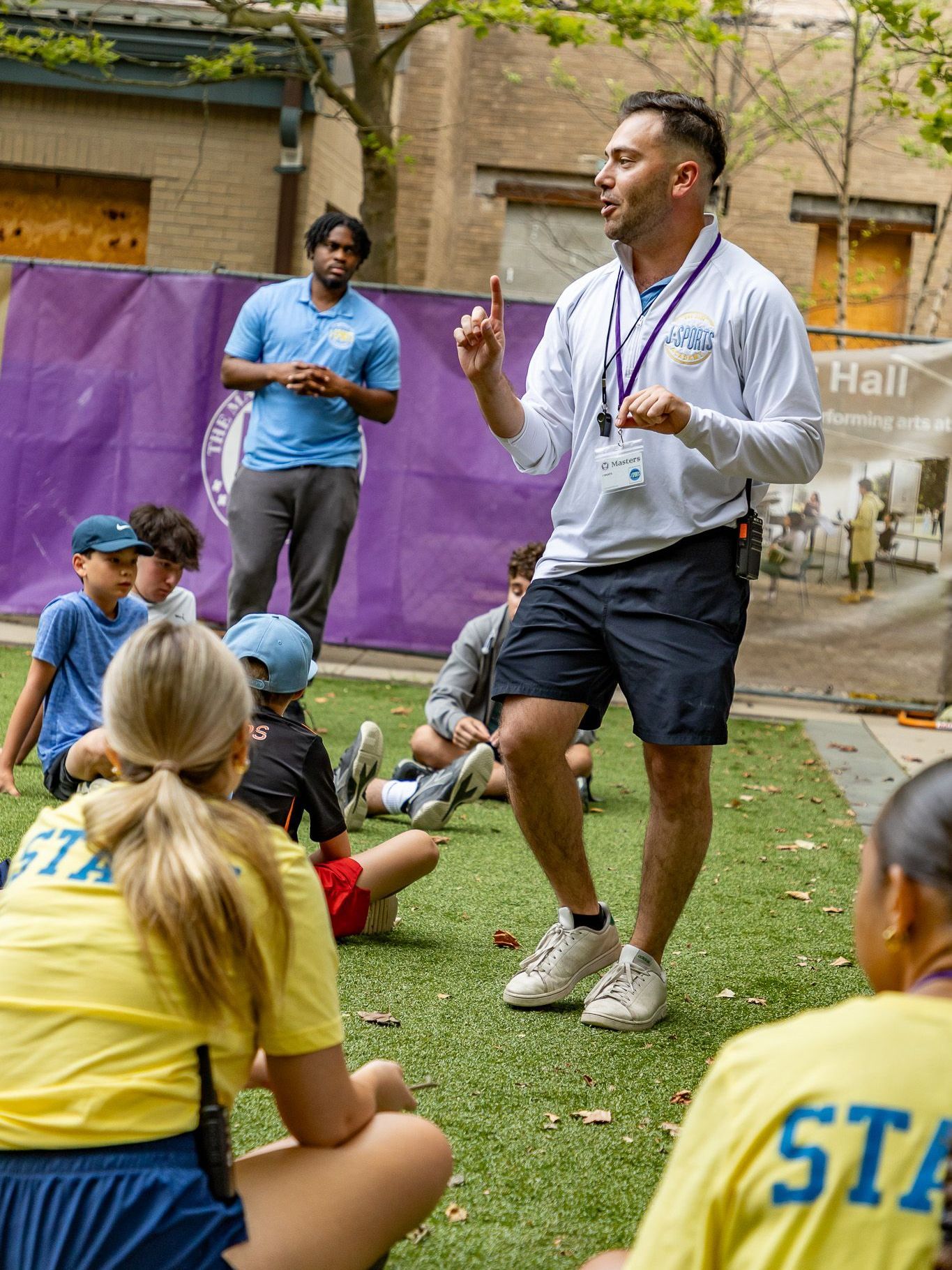 A man is standing in front of a group of children sitting on the grass.