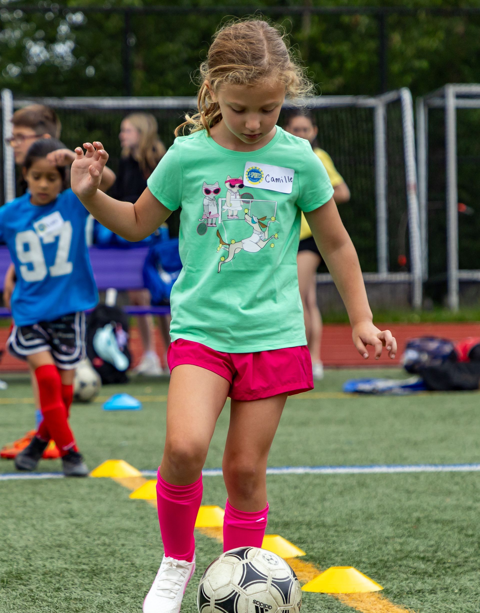 Girl in green shirt and pink shorts dribbles a soccer ball on a field. Other children and cones are in the background.